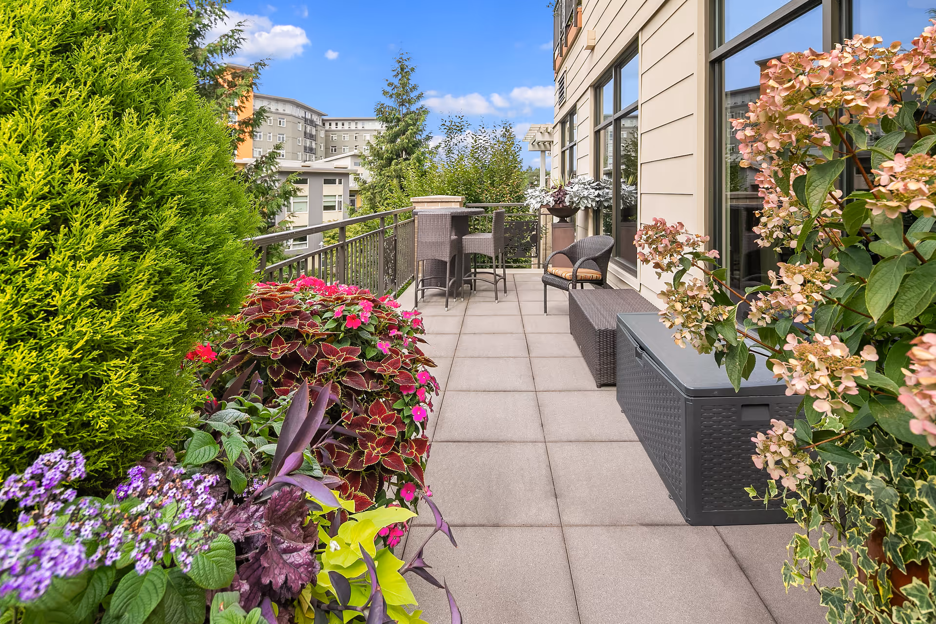 Outdoor patio area with tiled flooring, surrounded by lush green plants and colorful flowers. There are wicker chairs and a table near the railing, and a storage bench along the building wall with large windows reflecting the sky.