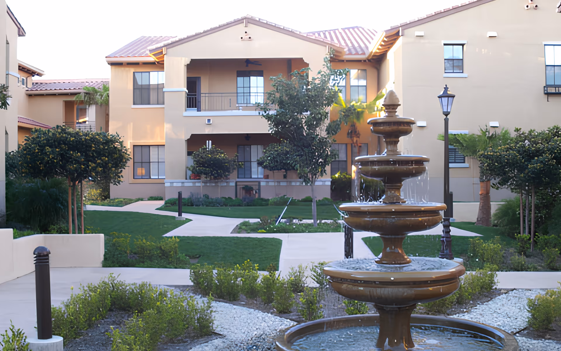 Tiered fountain in the courtyard of a two-story beige residential building with landscaped paths and greenery.