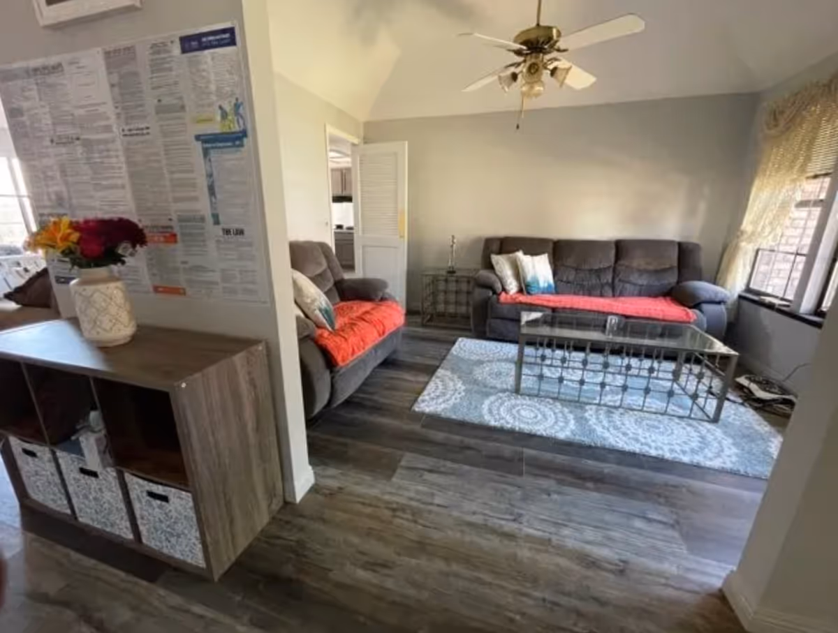 A cozy living room with two gray sofas covered with red throws and decorative pillows, a glass coffee table on a patterned rug, a ceiling fan, and a window with lace curtains. A wooden storage unit with baskets and a vase of flowers is visible in the foreground, along with a wall covered in informational posters.
