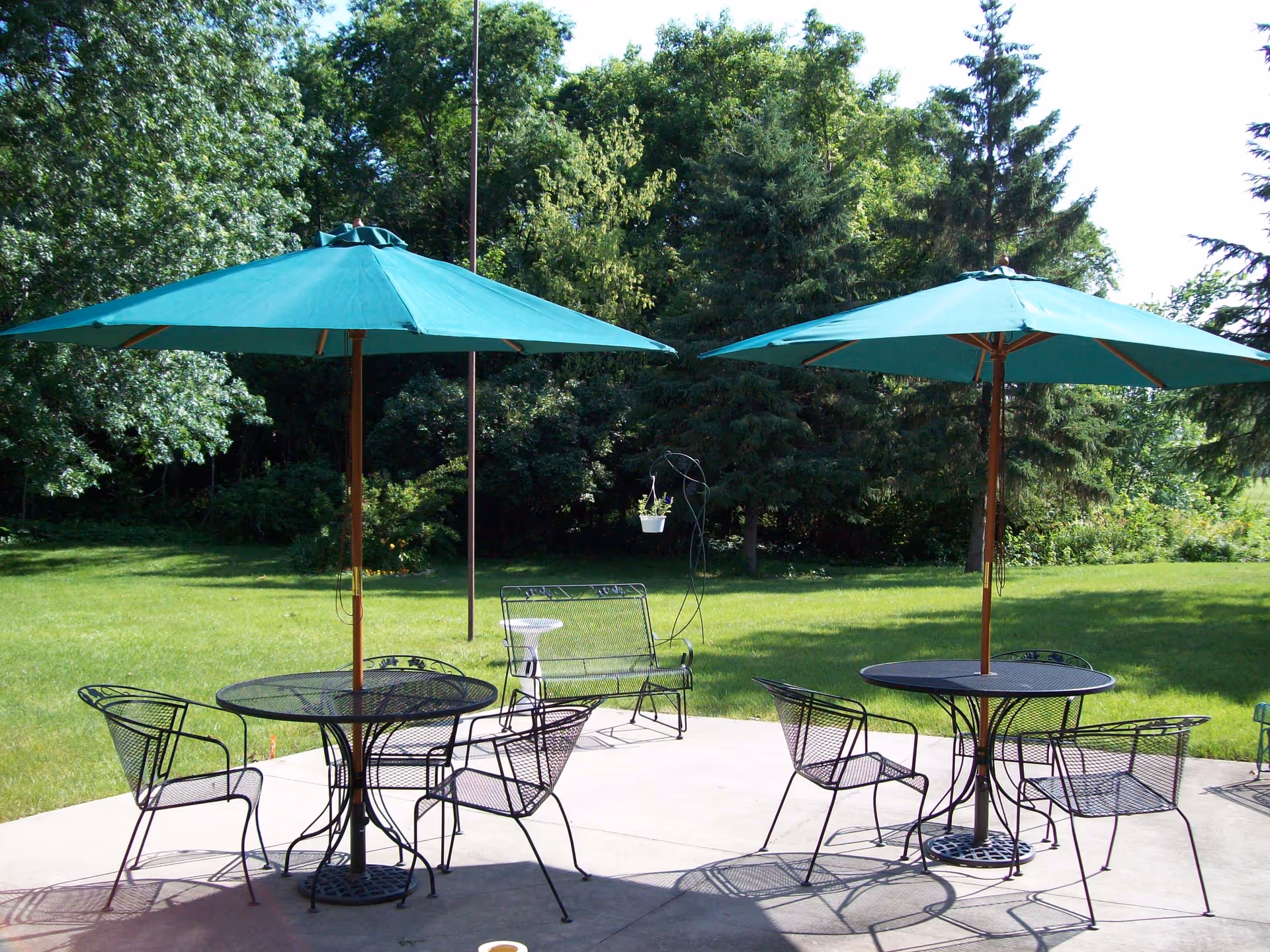 Outdoor patio area with two round metal tables, each with four matching metal chairs and a large green umbrella providing shade. The patio overlooks a grassy lawn with trees and shrubs in the background.