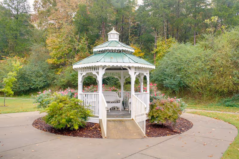 A white wooden gazebo with a green roof situated in a garden area surrounded by bushes and flowers, with a paved walkway encircling it and trees in the background.