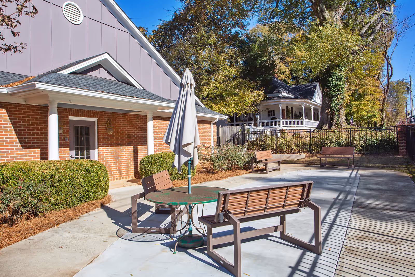 Outdoor patio area at Bethel Gardens Assisted & Memory Care with benches, a round table with a closed umbrella, surrounded by bushes and trees, adjacent to a brick building.