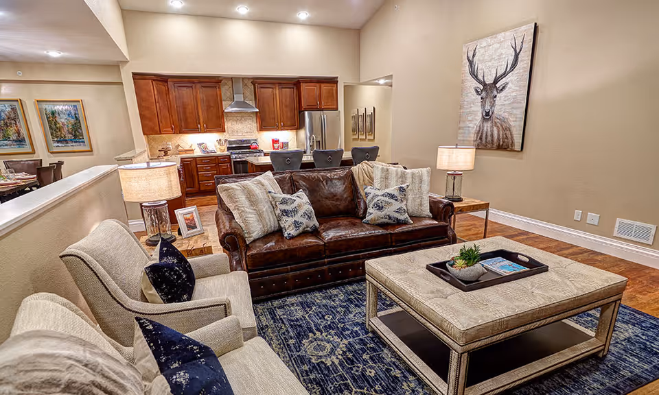 A cozy living room area with a brown leather sofa adorned with multiple pillows, two beige armchairs with dark blue pillows, a large beige ottoman coffee table with a tray holding a small plant and magazines, and two side tables with lamps. The room has wooden flooring and a blue patterned rug. In the background, there is a kitchen with wooden cabinets, a stainless steel refrigerator, and a stove. A large painting of a deer hangs on the wall.
