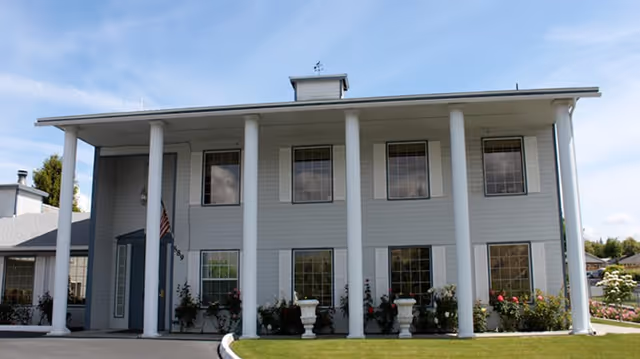 Front exterior view of a two-story senior living facility building with six large white columns, multiple windows with white shutters, and a well-maintained lawn with flower beds in front.