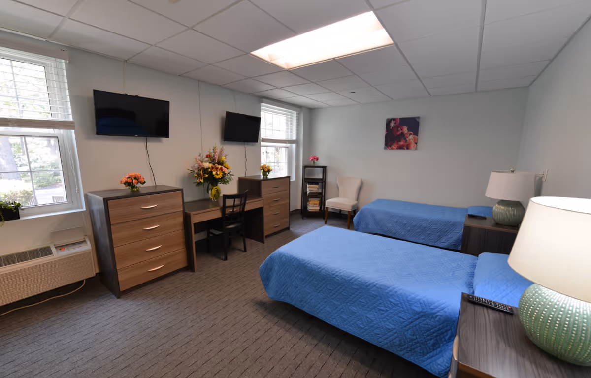 Twin bedroom with two blue-covered beds, dressers, wall-mounted TVs, a desk, and seating.