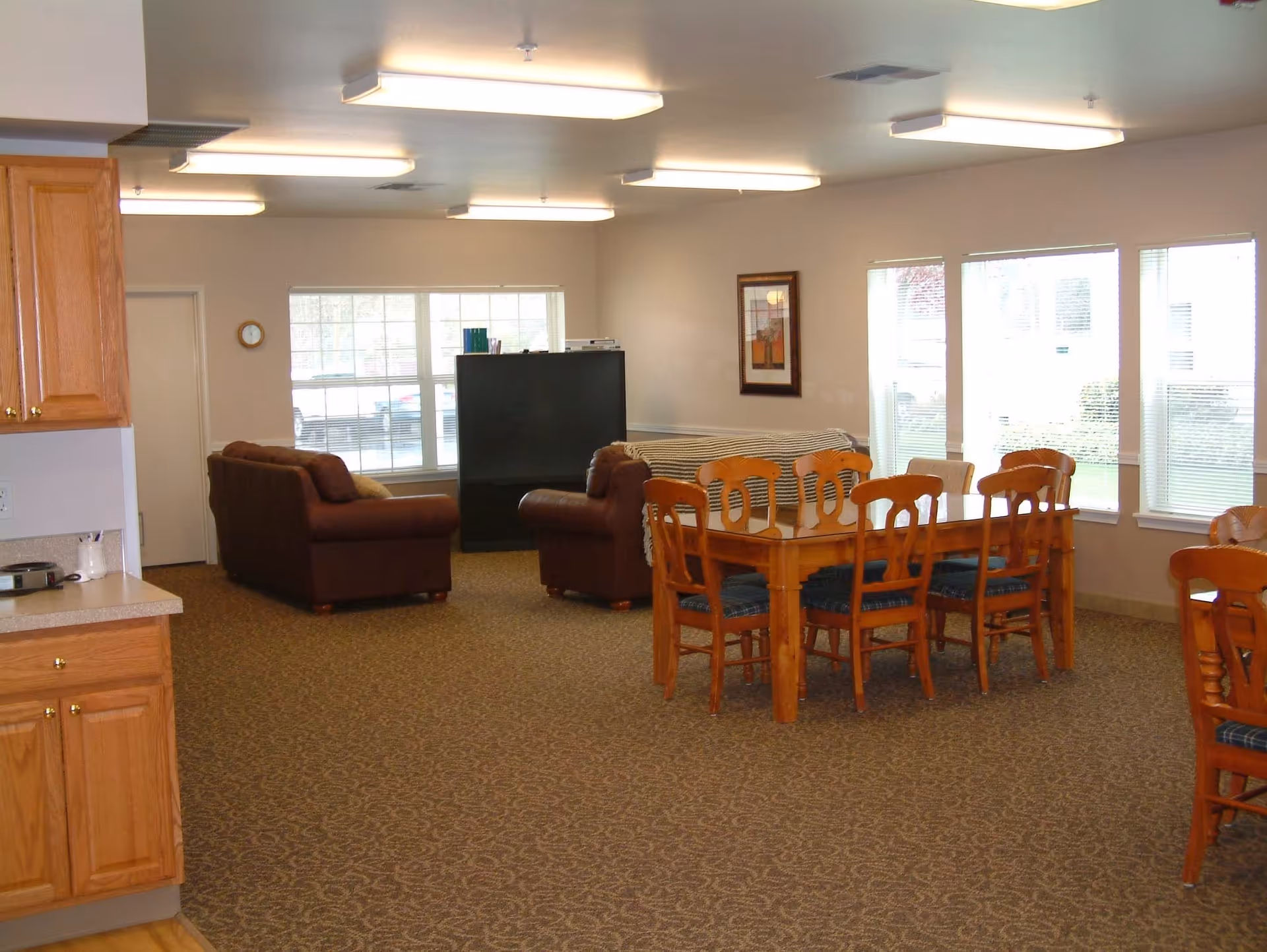 A spacious room with a dining area featuring a wooden table and six chairs with blue cushions, a living area with two brown sofas and a TV, large windows letting in natural light, and part of a kitchen with wooden cabinets visible on the left.