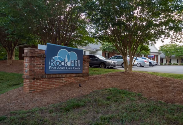 Outdoor view of the Rock Hill Post Acute Care Center sign mounted on a brick base, with several parked cars and trees surrounding the area.