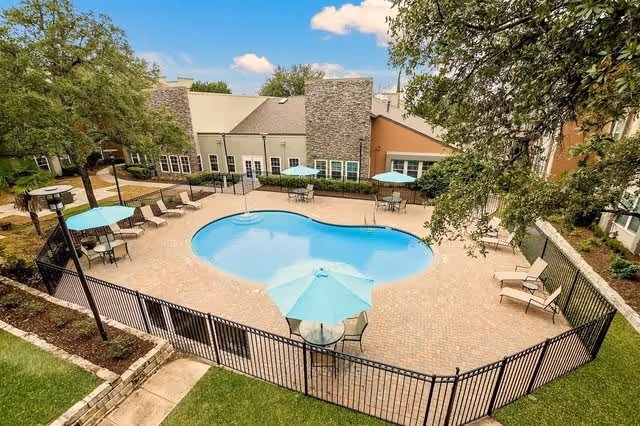 Outdoor swimming pool area at New Forest Haven with a kidney-shaped pool surrounded by a brick patio. Several tables with blue umbrellas and lounge chairs are arranged around the pool. The area is fenced and bordered by trees and a building in the background under a partly cloudy sky.