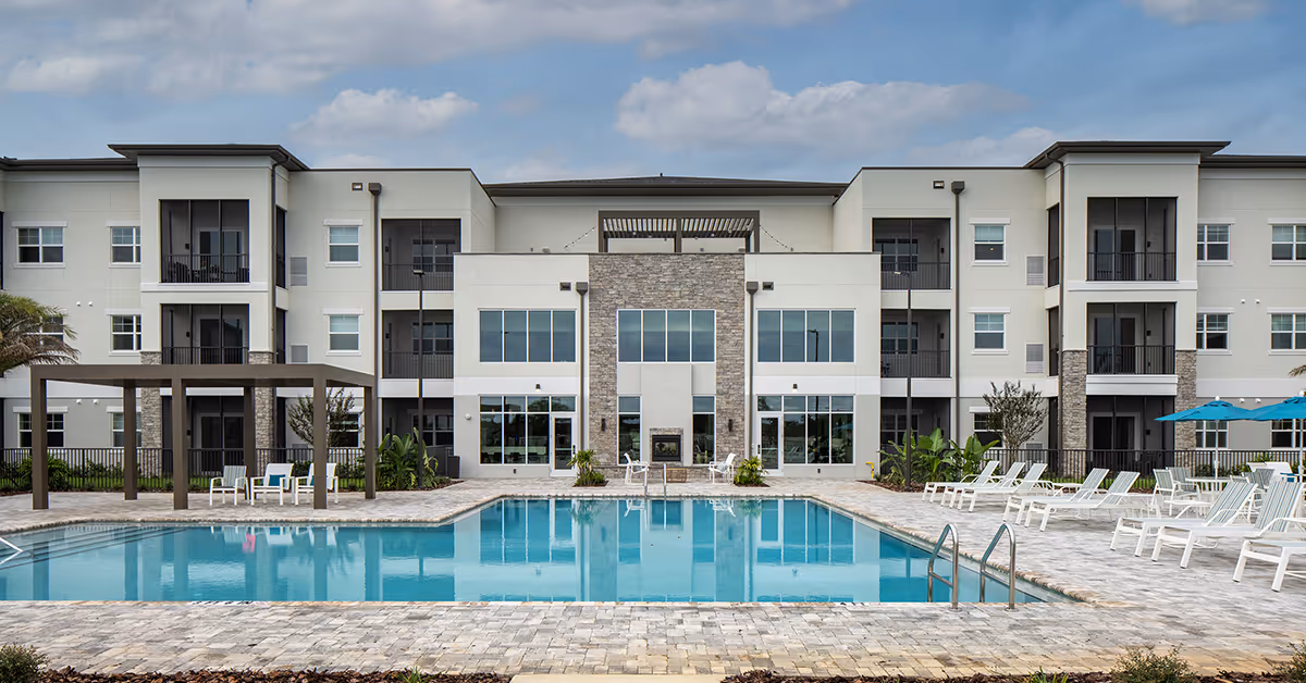 Outdoor swimming pool with lounge chairs and umbrellas in front of a modern three-story residential building under a partly cloudy sky.