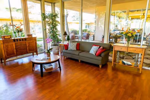 Cozy senior living lounge with sofa, coffee table, plants, and large windows.