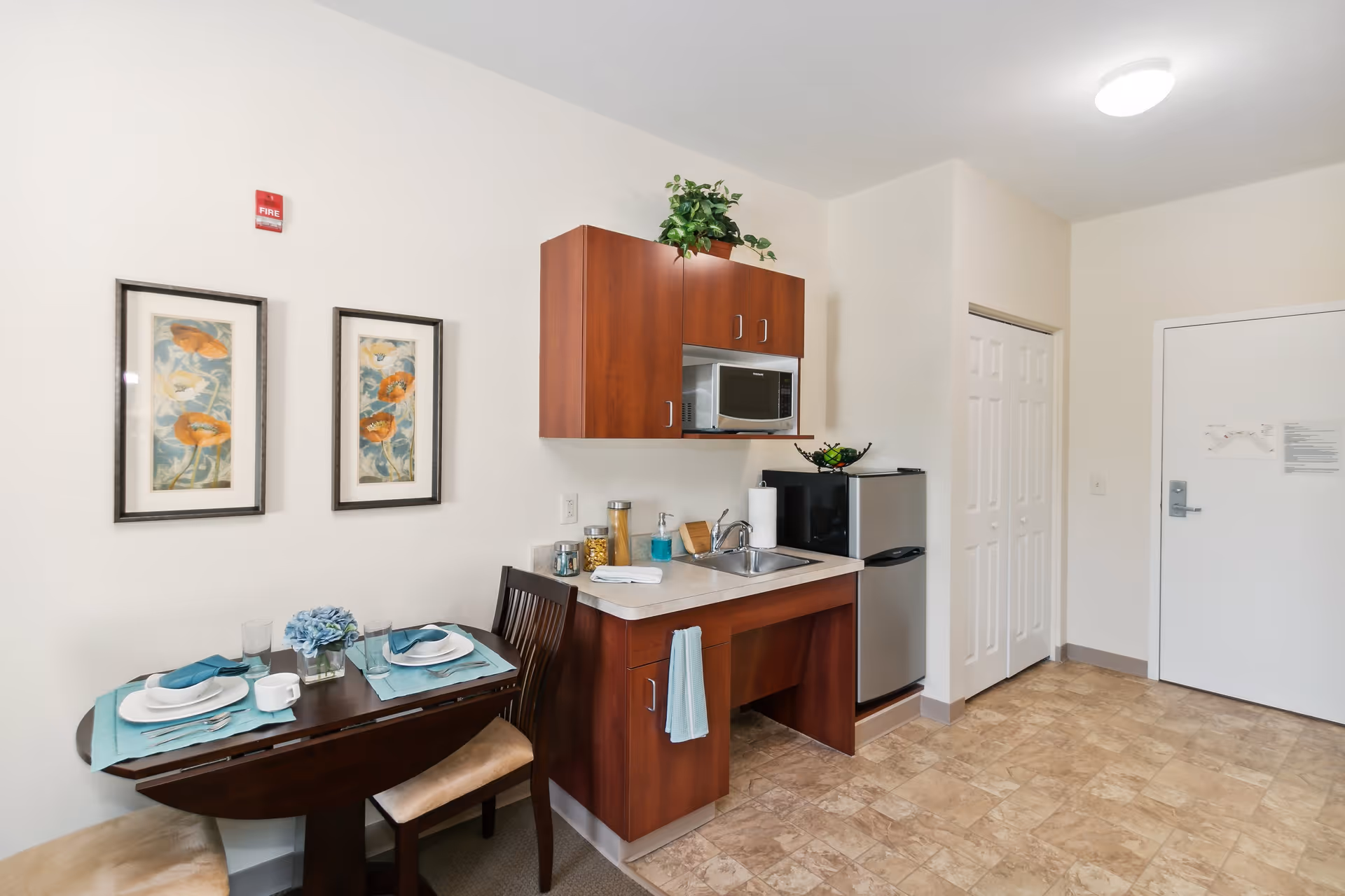 A small kitchenette area with a wooden table set for two with plates, glasses, and napkins. Above the table are two framed floral paintings. The kitchenette includes a sink, a microwave mounted above, a small refrigerator, and wooden cabinets. The floor is tiled, and there is a white door and a closet door in the background.