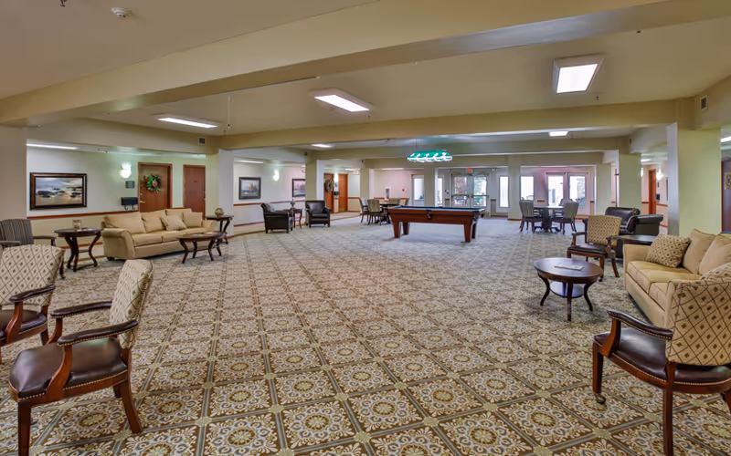 A spacious senior living facility common area with patterned carpet, multiple seating arrangements including sofas and armchairs, a pool table in the center, and large windows letting in natural light.