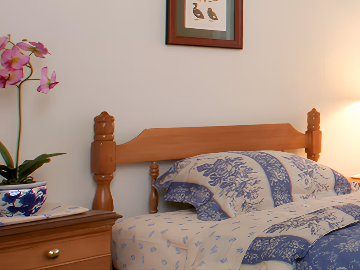 Wood-framed bed with blue floral bedding, a wooden nightstand holding a potted orchid and lamp, and a framed picture on the wall.