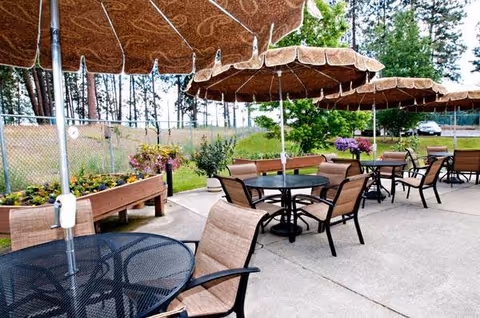 Outdoor patio area with several round tables and cushioned chairs under large patterned umbrellas. Raised garden beds with flowers and plants line the fence in the background, with trees and greenery beyond the fence.