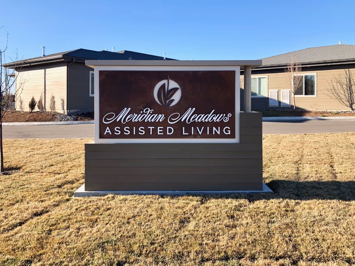 Outdoor sign for Meridian Meadows Assisted Living on a grassy area with buildings in the background under a clear blue sky.