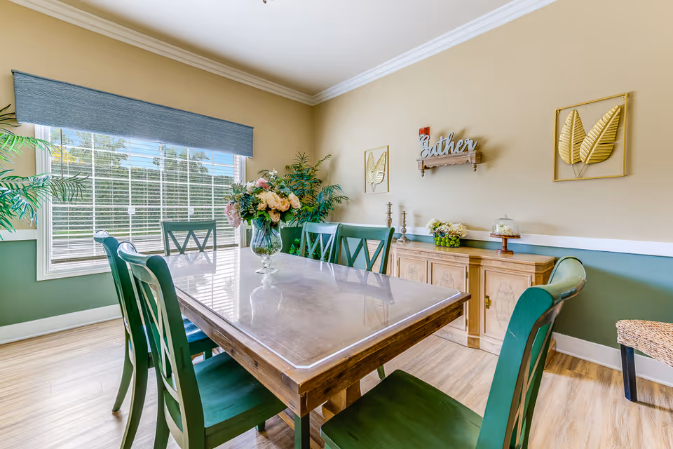 A bright dining room with a wooden table covered by a clear protective surface and surrounded by green chairs. A large window with blinds and a blue valance lets in natural light. The walls are painted beige and green with white trim, decorated with gold leaf wall art and a shelf with the word 'Gather'. A wooden sideboard holds decorative items including flowers and candlesticks.