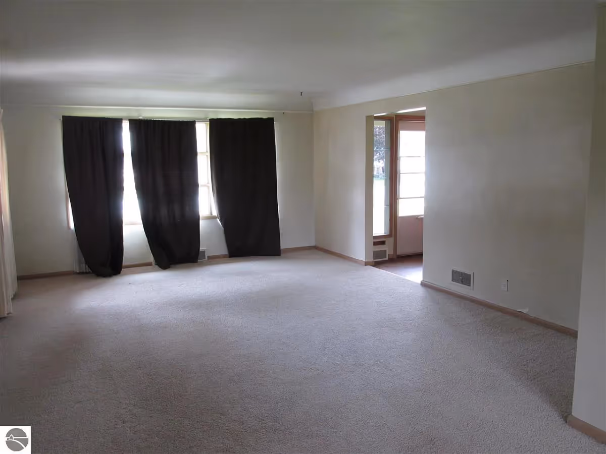 Empty living room with beige carpet, cream-colored walls, three windows covered with dark curtains, and an open doorway leading to another room with a glass door.