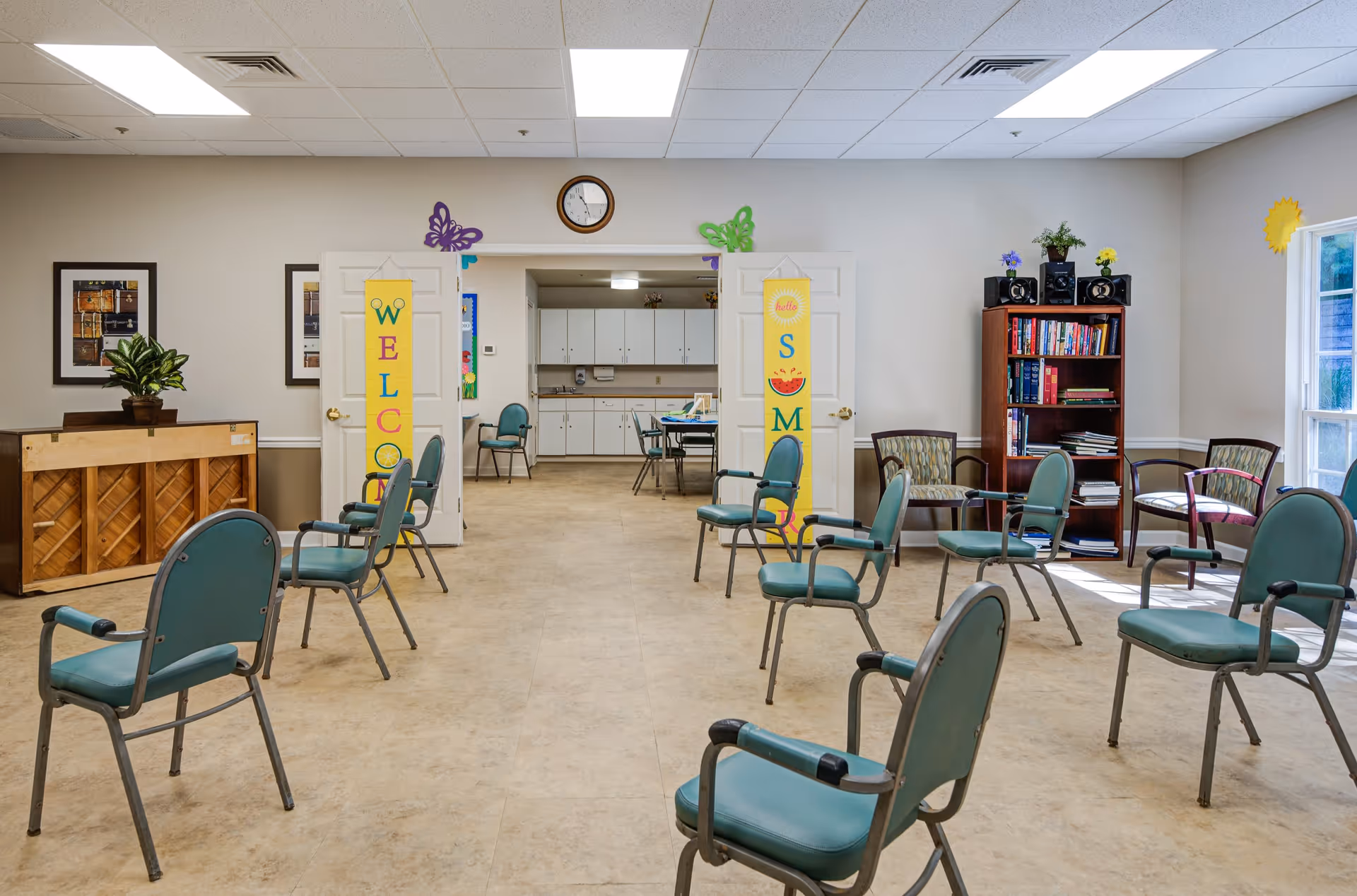 A spacious room with chairs arranged in a circle, a piano on the left side, and a bookshelf with books and speakers on the right. The room has a tiled floor, white walls, and a clock above double doors that lead to another room with tables and chairs. Colorful welcome banners hang on the doors, and there are windows letting in natural light.