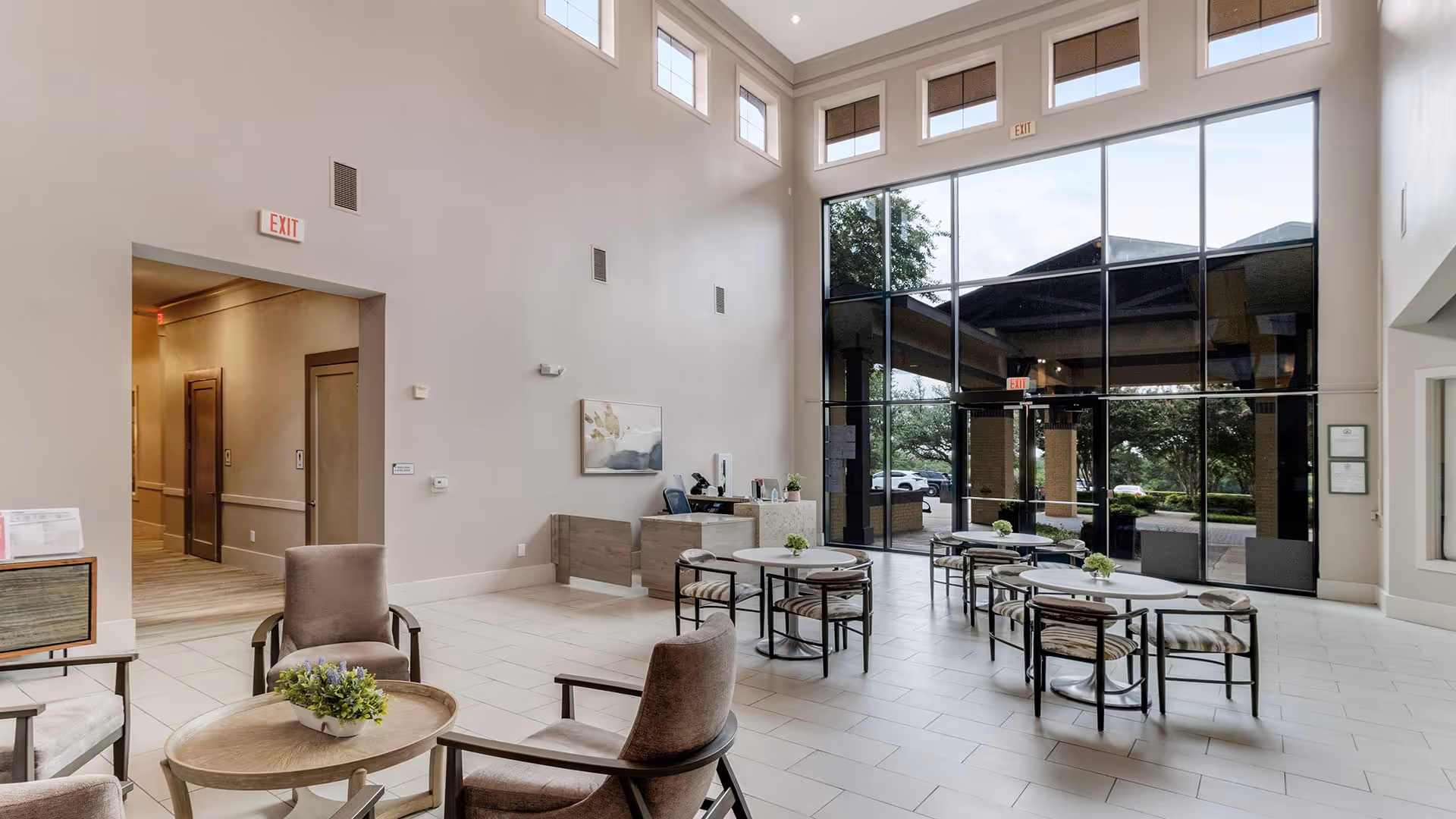 A spacious and bright common area in a senior living facility with high ceilings and large floor-to-ceiling windows letting in natural light. The room features several round tables with chairs and small green plants on each table. There are also comfortable armchairs around a small round coffee table with a plant centerpiece. The walls are painted light beige, and there is a reception desk in the background near the entrance. The floor is tiled in a light color, and there are exit signs above doorways.
