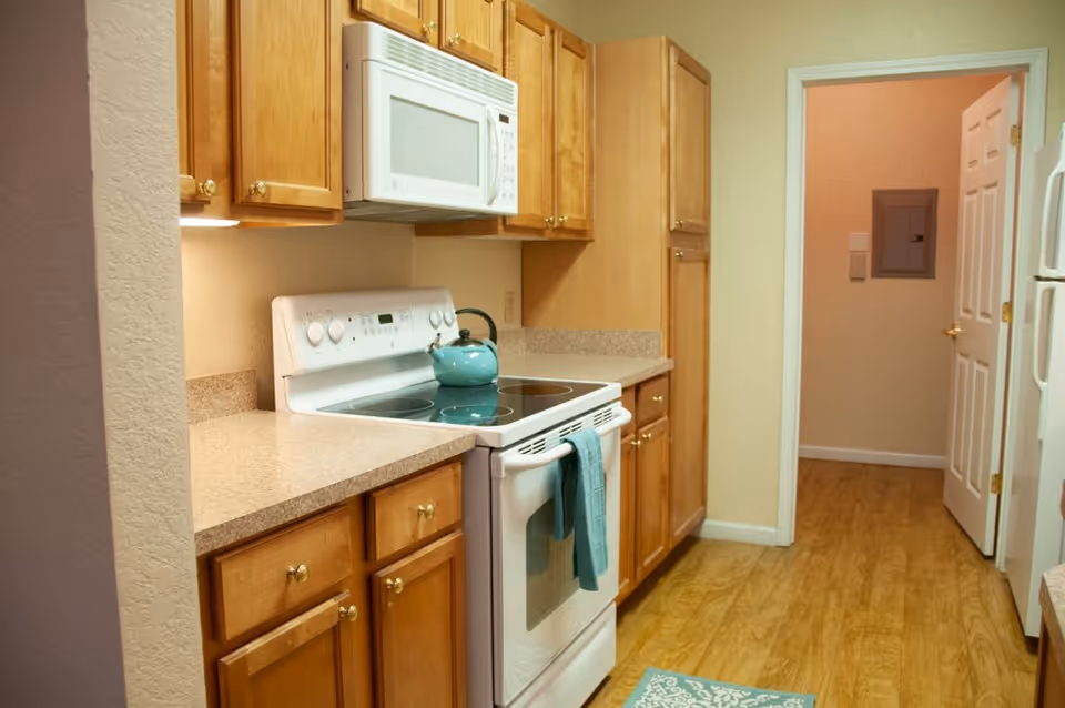 Small kitchen with wooden cabinets, a white stove and microwave, a teal kettle and towel, and wood-look flooring.