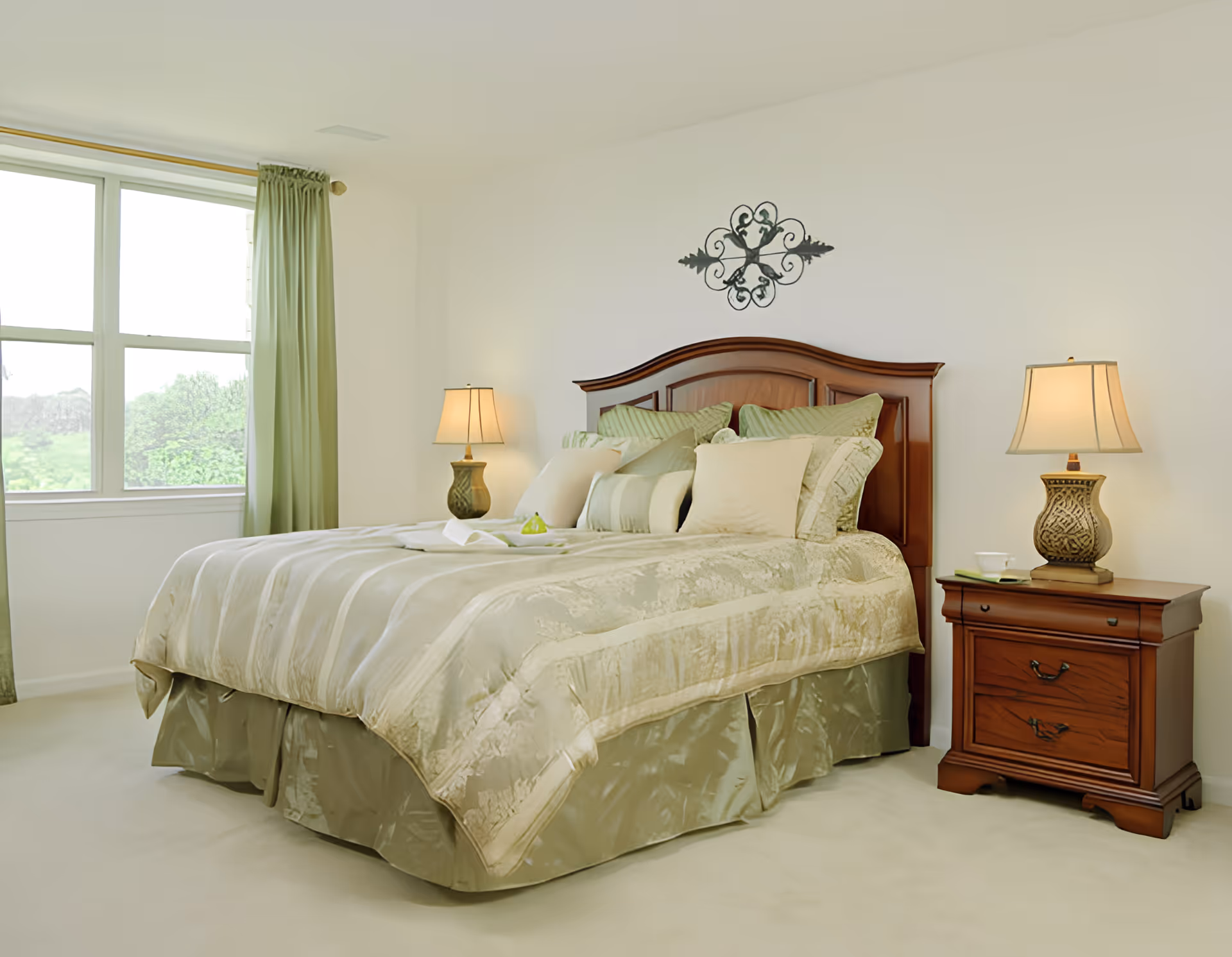 A neatly made bed with a wooden headboard in a bright bedroom. The bed has multiple pillows and a light green and beige comforter. There are two matching bedside tables with decorative lamps on each side of the bed. A window with green curtains lets in natural light, and a decorative metal wall art piece hangs above the headboard.
