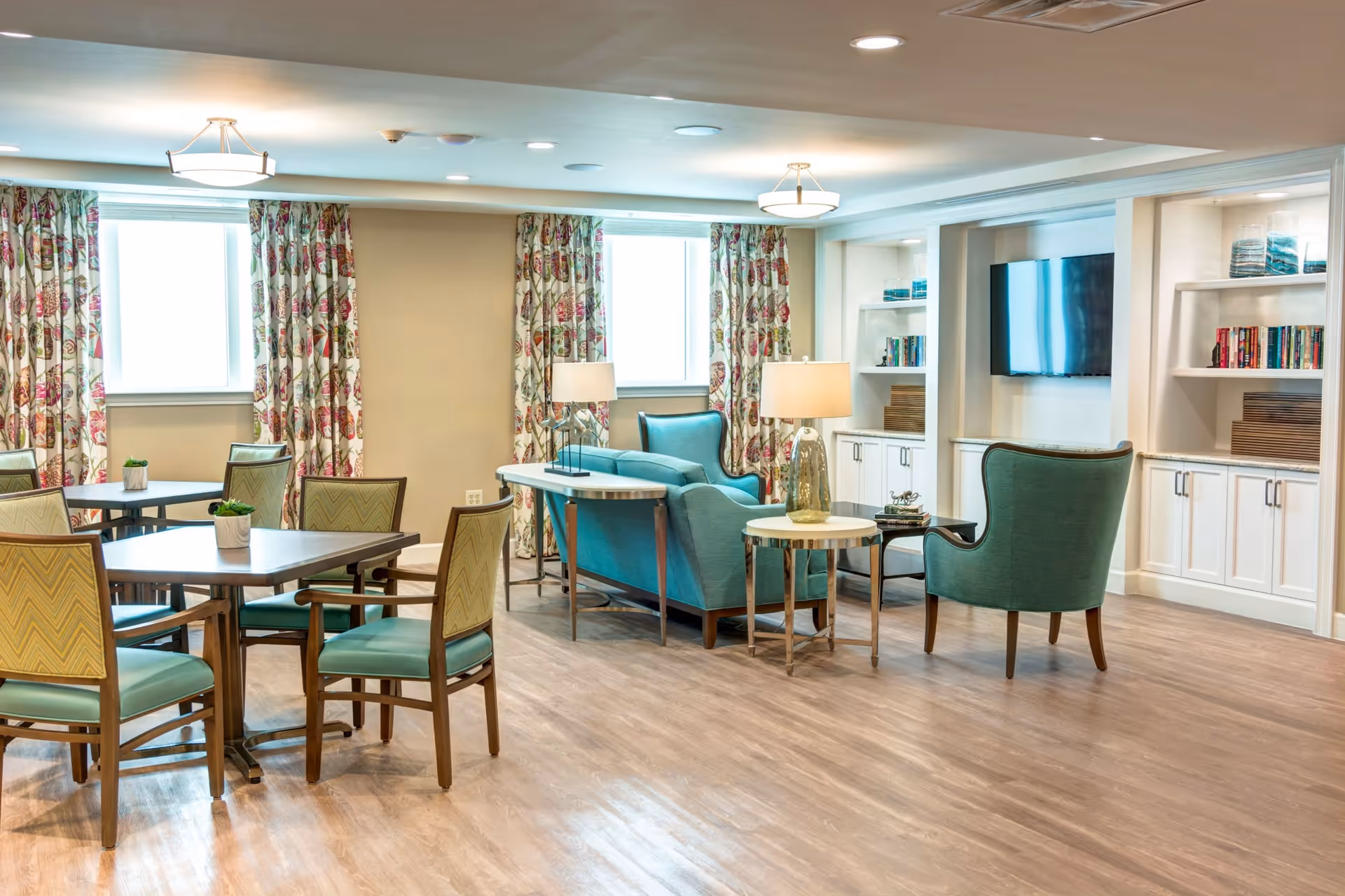 A bright and spacious common area in a senior living facility featuring multiple tables with chairs, a seating area with teal upholstered armchairs and a sofa, side tables with lamps, built-in white shelves with books and decorative items, and a wall-mounted flat-screen TV. The room has large windows with floral curtains and wood flooring.