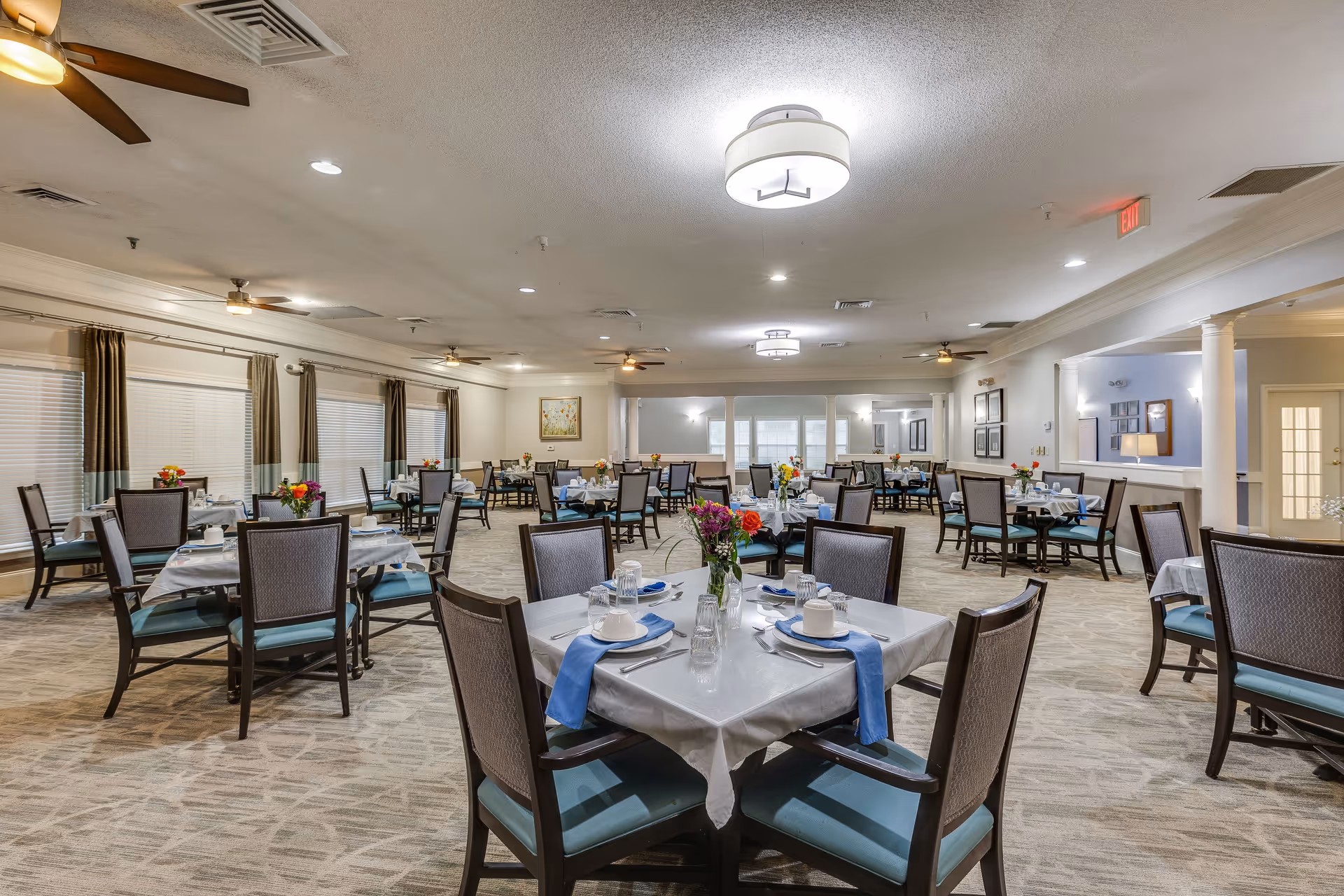 A spacious dining room in a senior living facility with multiple tables set for meals. Each table has a white tablecloth, blue napkins, plates, cups, and a vase with colorful flowers. The room is well-lit with ceiling lights and ceiling fans, and windows with curtains line one side of the room.