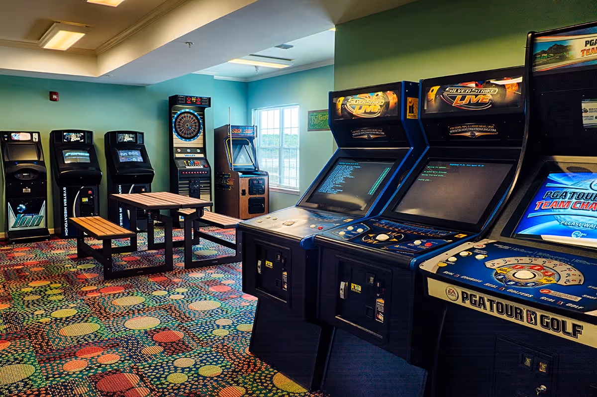 A colorful game room with multiple arcade machines, including Silver Strike Live and PGA Tour Golf, and a dartboard on the far wall. There are two wooden picnic-style tables with benches in the center of the room. The walls are painted light blue and there is a window letting in natural light.