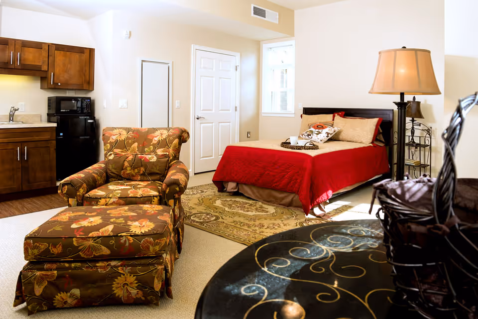 A cozy studio apartment featuring a bed with a red blanket and decorative pillows, a floral-patterned armchair with matching ottoman, a small kitchenette with wooden cabinets, a black microwave and mini fridge, a floor lamp, and a round table with a decorative metal basket. The room has beige walls and carpet, with a window letting in natural light.
