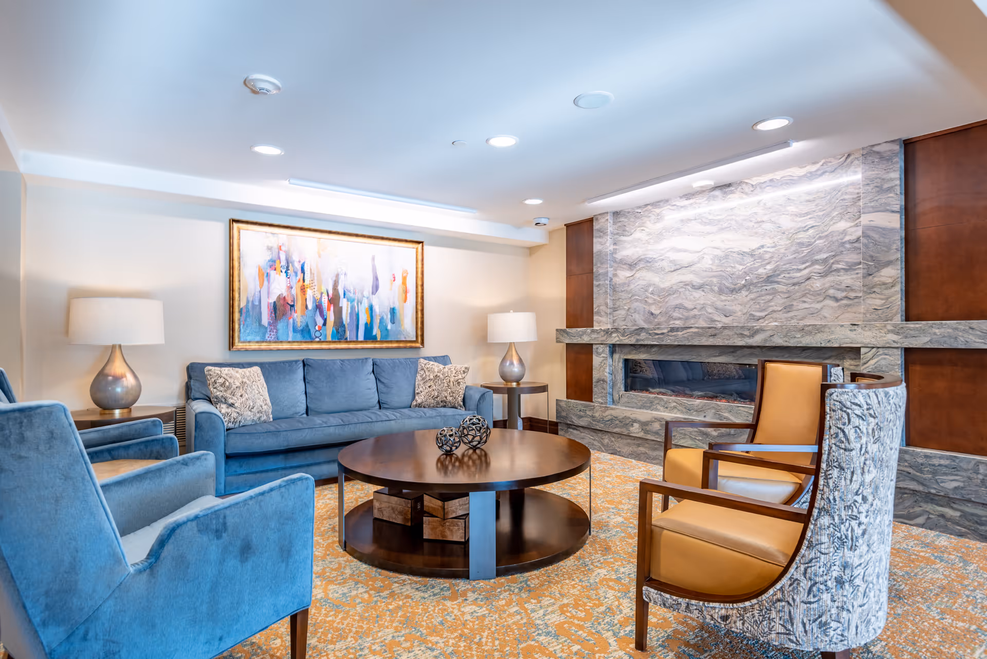 A cozy living room in Frasier Retirement Community featuring a blue sofa with patterned pillows, two blue armchairs, and two patterned armchairs with tan cushions arranged around a round wooden coffee table. The room has a modern stone fireplace with a marble-like finish, two table lamps with beige shades, and a colorful abstract painting on the wall above the sofa. The carpet has a warm orange and beige pattern.