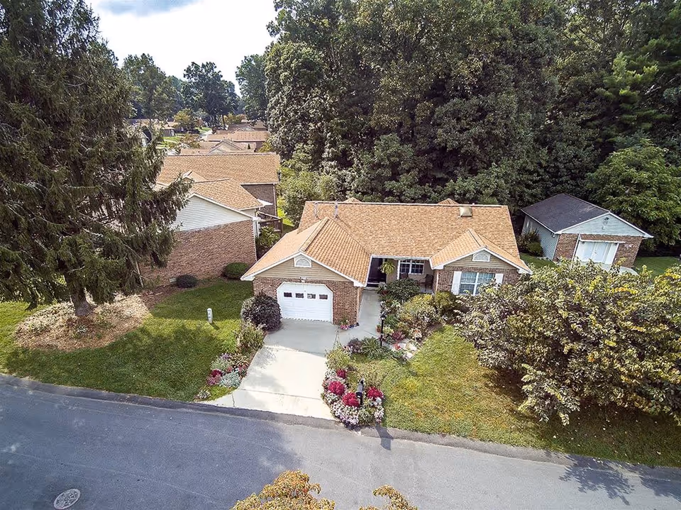 Aerial view of a single-story brick house with a tan shingle roof, a driveway leading to a white garage door, and a well-maintained front yard with green grass, colorful flower beds, and surrounding trees. Additional similar houses and dense trees are visible in the background.
