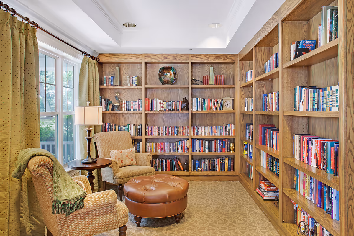 Cozy reading room with built-in wooden bookshelves, two upholstered armchairs, a leather ottoman, and a window with green curtains.