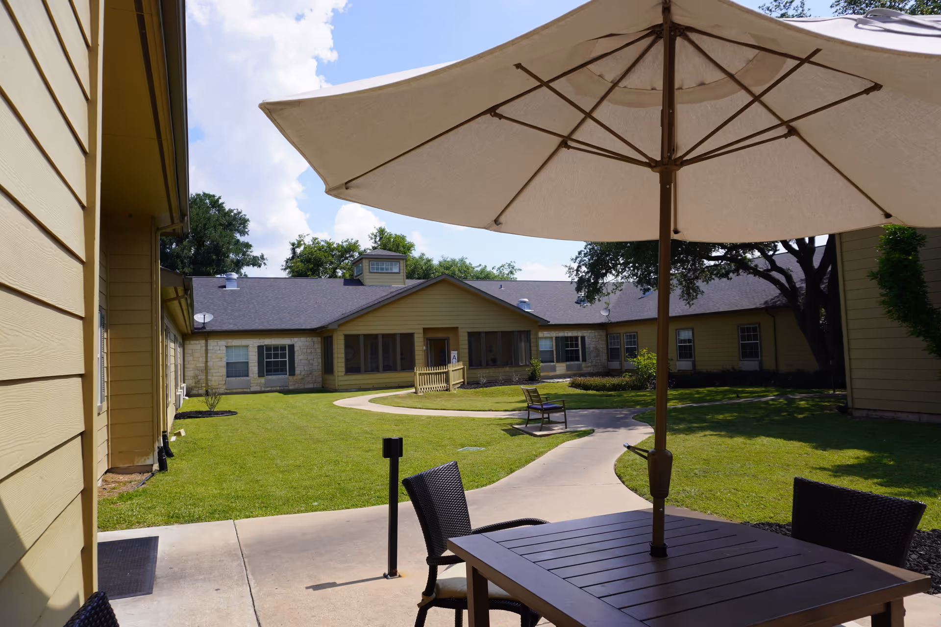 Outdoor courtyard area at Sodalis Austin featuring a patio table with an umbrella and chairs, a winding concrete pathway, green grass, benches, and yellow buildings with stone accents under a partly cloudy sky.