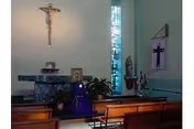 Interior of a small chapel with wooden pews, a crucifix mounted on the wall above an altar, a purple cloth draped over the altar, religious statues, and a stained glass window allowing light into the space.