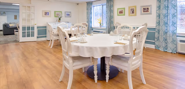 A bright dining room with a round table covered with a white tablecloth and set with glasses, napkins, and utensils. The room has wooden flooring, white chairs around the table, large windows with blue patterned curtains, and framed pictures on the walls. A doorway leads to another room with a sofa visible.