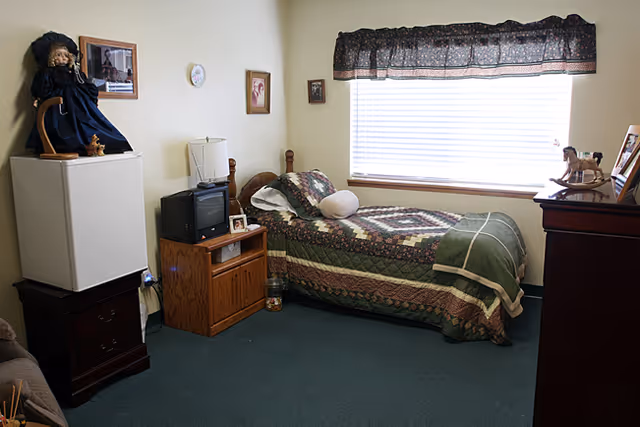 A small bedroom with a single bed covered in a green and brown patchwork quilt. The room has a window with blinds and a dark valance. There is a wooden nightstand with a small TV and a lamp, a white mini fridge with a doll on top, and a wooden dresser with framed photos and a small rocking horse figurine. The walls have a few framed pictures and a clock.