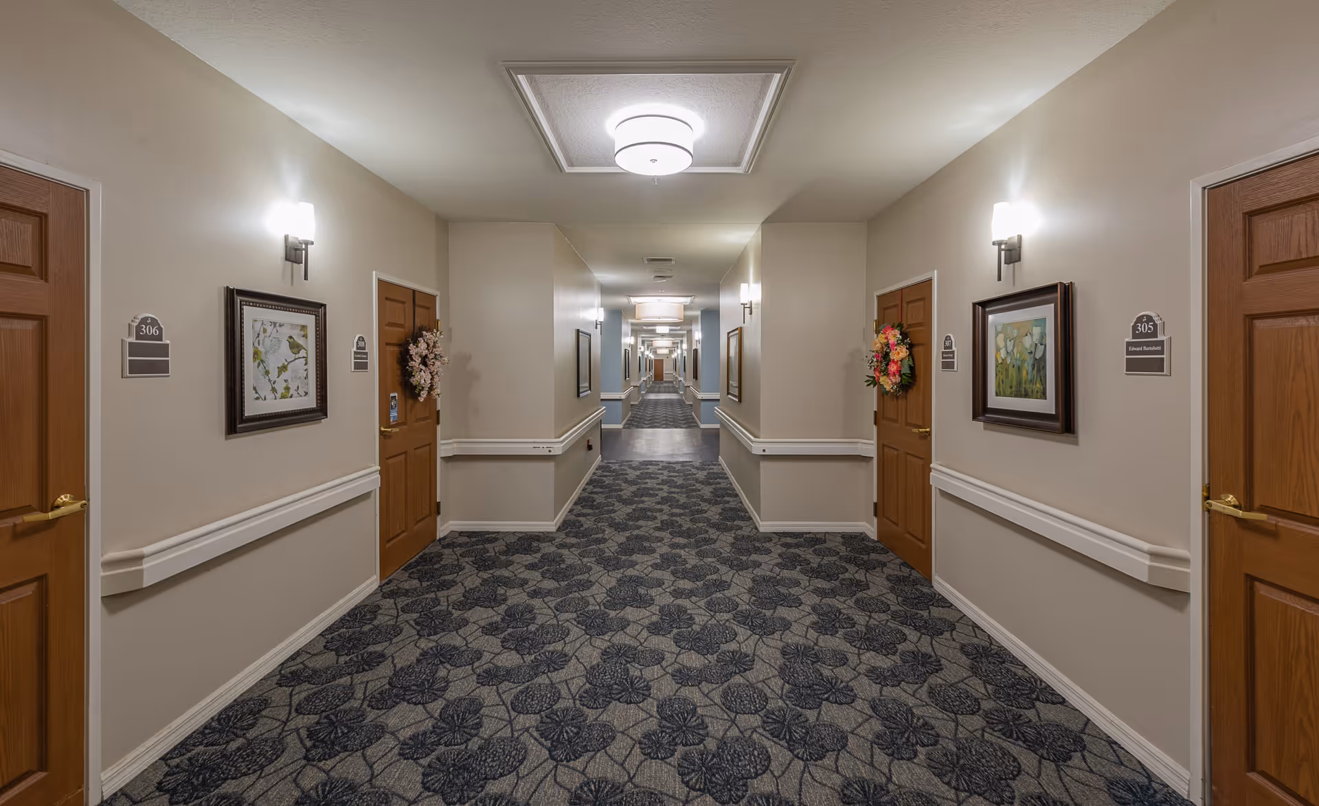 A long, well-lit hallway in a senior living facility with beige walls, patterned carpet, and wooden doors on both sides. Each door has a room number and some have decorative wreaths. Framed artwork hangs on the walls between the doors, and handrails run along both sides of the hallway.