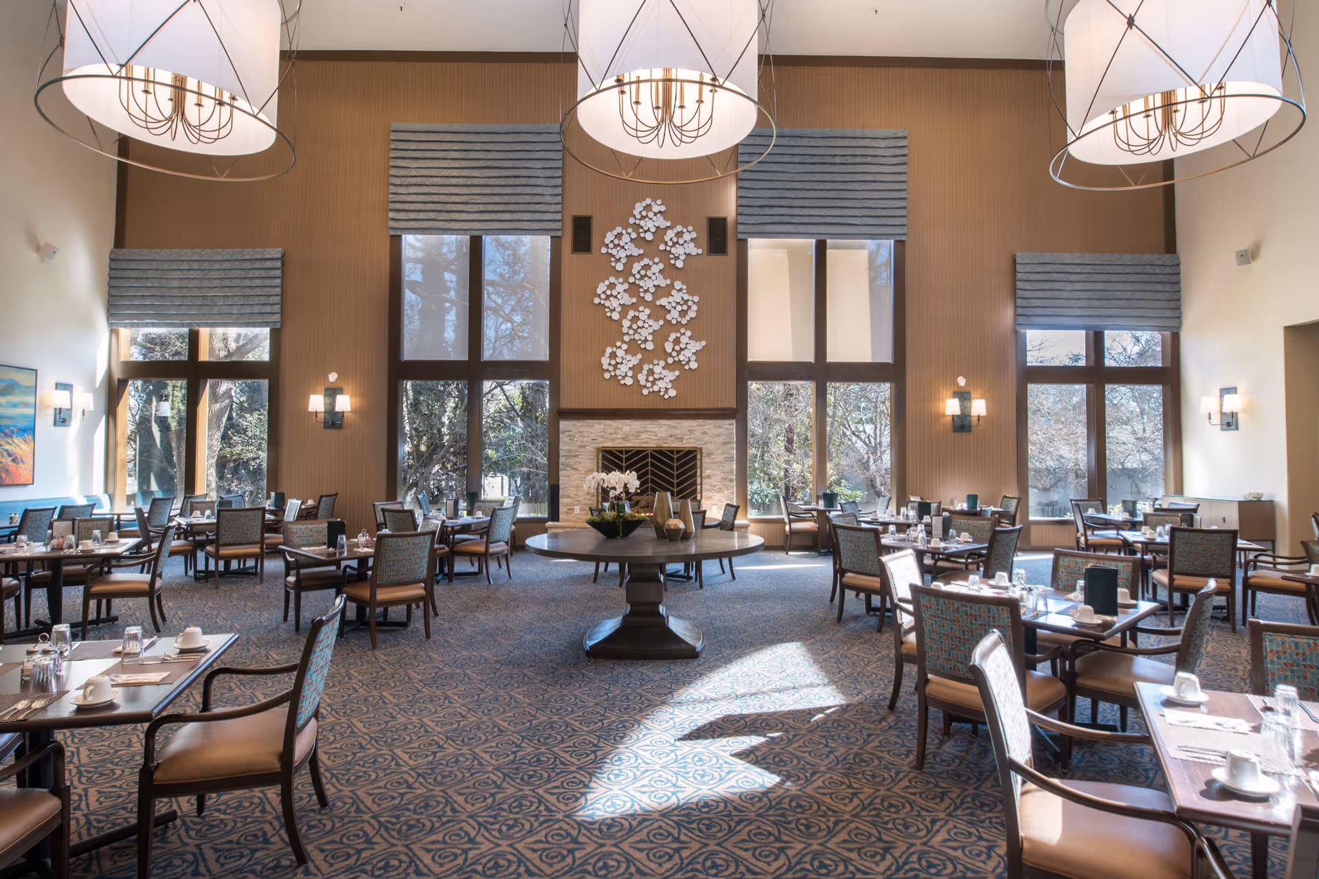 A spacious dining room with multiple tables and chairs arranged neatly. The room features large windows with blue Roman shades, allowing natural light to fill the space. There is a central round table with decorative items and a fireplace with a floral wall decoration above it. Three large pendant lights hang from the ceiling, and the carpet has a patterned design.