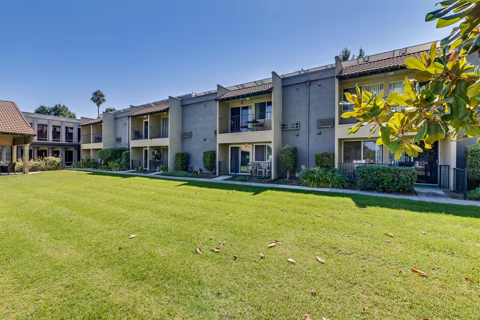 Exterior view of a two-story senior living facility building with balconies and patios overlooking a well-maintained grassy lawn under a clear blue sky. There are shrubs and trees around the building and a palm tree in the background.