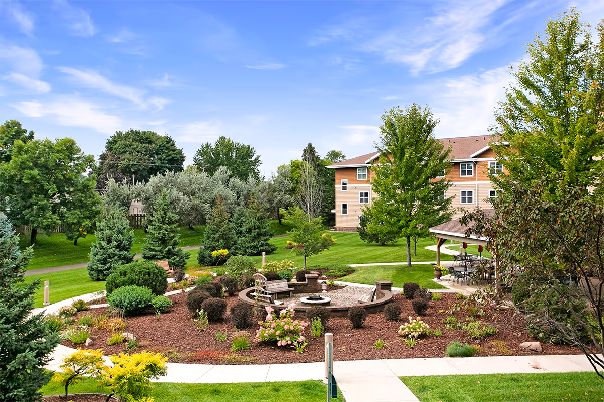 Landscaped courtyard with a circular fountain, benches, walking paths and trees in front of a multi-story residential building.