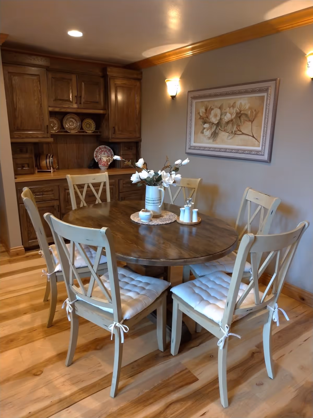 Cozy dining room with a round wooden table surrounded by six cream chairs with cushions, a floral centerpiece, wall art, and built-in wooden cabinets.