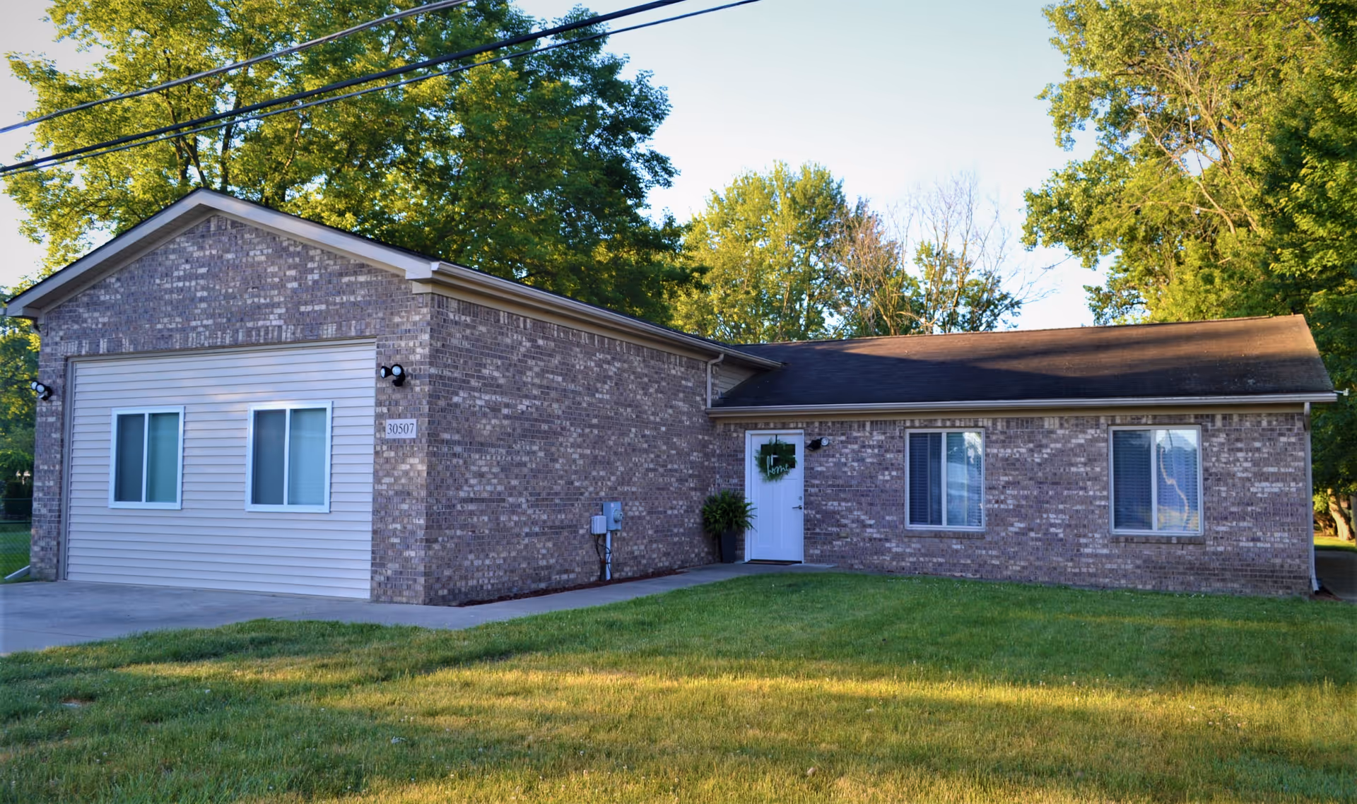 Exterior view of a single-story brick building with a white door decorated with a wreath, three windows, and a driveway. The building is surrounded by green grass and trees under a clear blue sky.