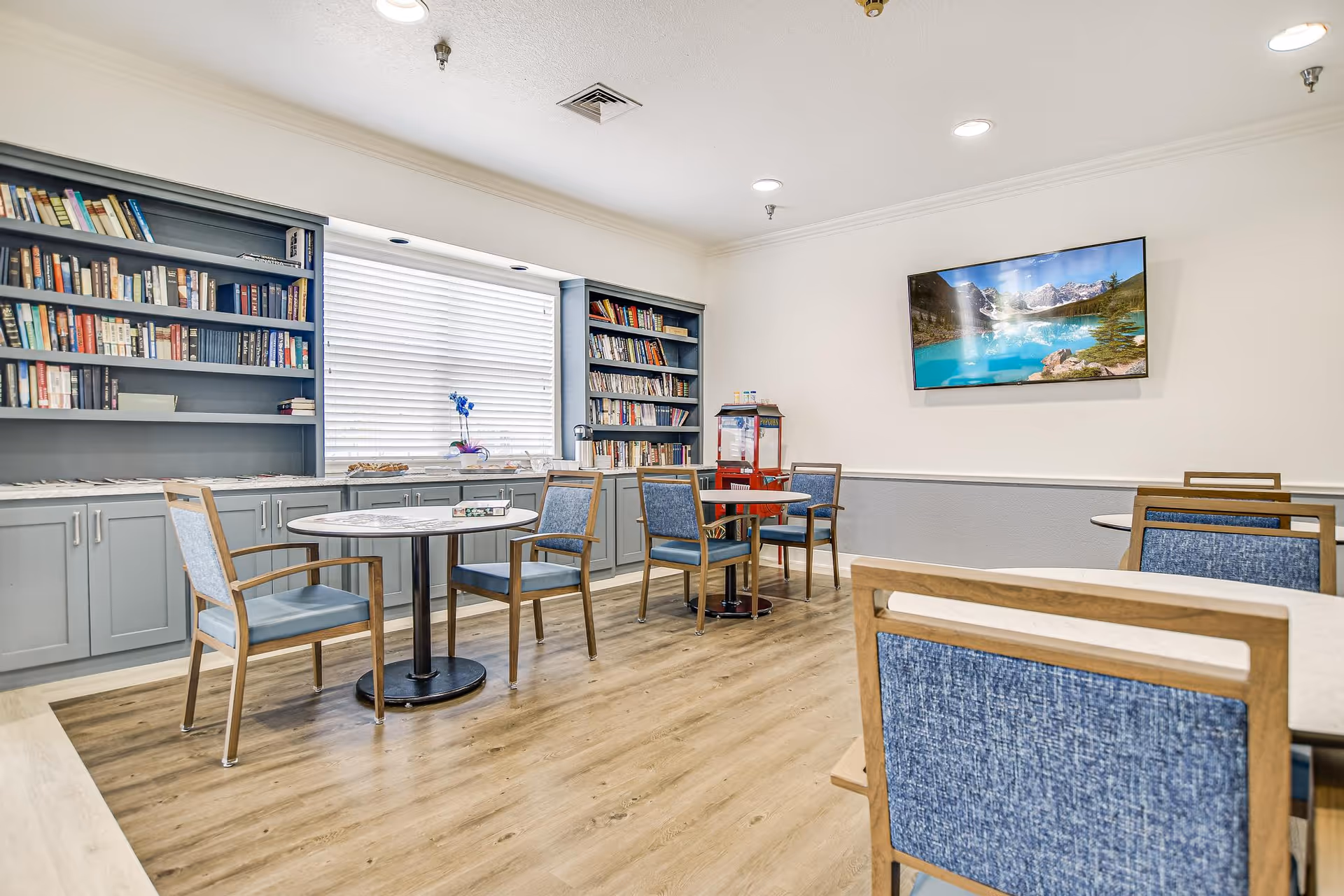 A bright room with wooden flooring featuring several round tables and blue cushioned chairs. Built-in bookshelves filled with books line one wall, with a window in the center letting in natural light. A flat-screen TV mounted on the opposite wall displays a scenic mountain and lake image. A popcorn machine is placed near the corner.