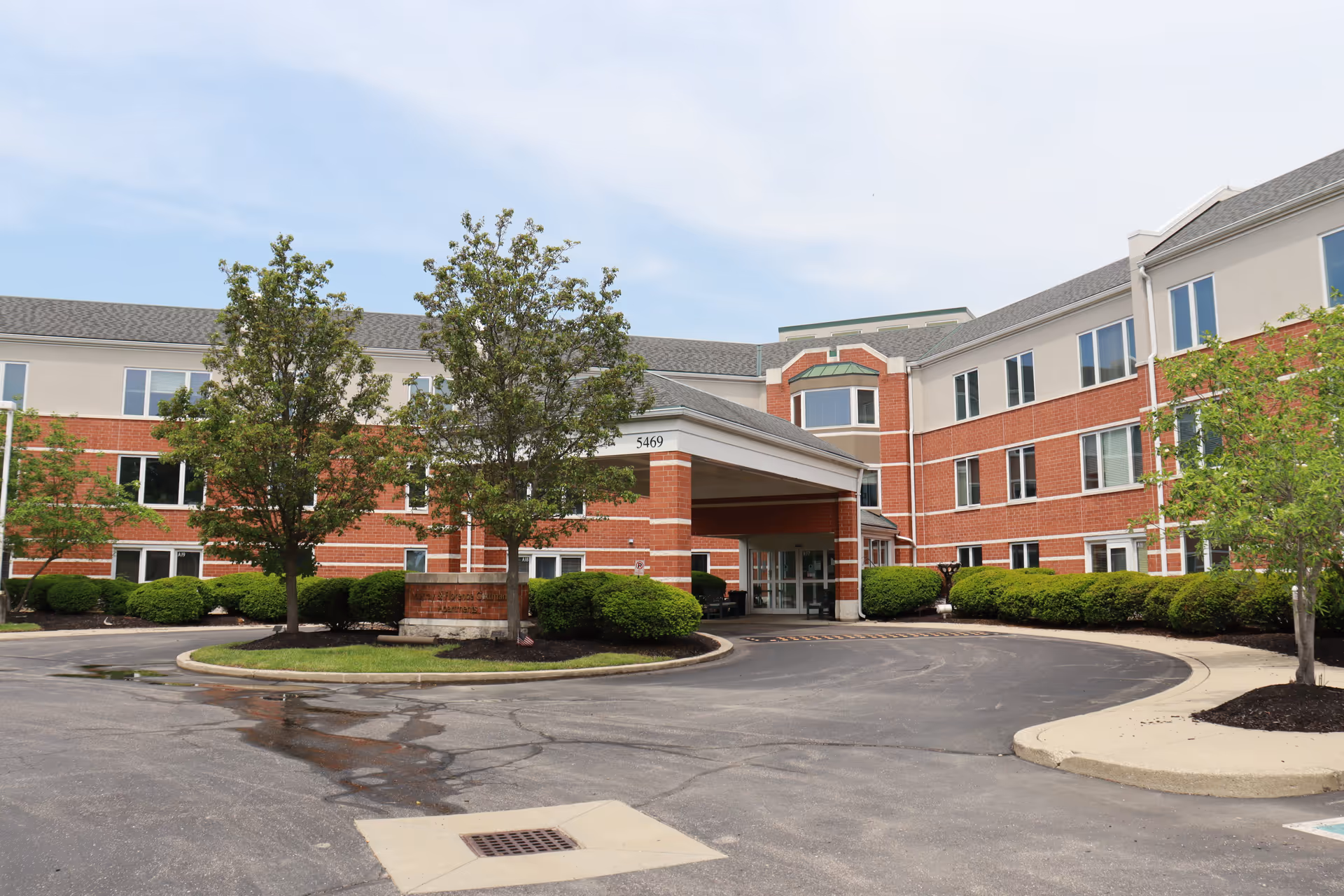 Front exterior view of a three-story senior living facility building with a covered entrance, surrounded by trees and bushes, under a partly cloudy sky.