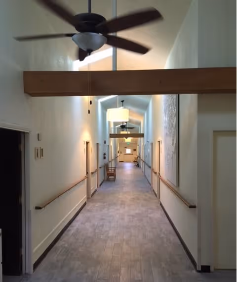 A long, well-lit hallway with light-colored walls and wooden handrails on both sides. The ceiling has exposed wooden beams and ceiling fans with lights. There are several doors along the hallway and a chair placed against the wall. The floor is covered with light-colored tiles.