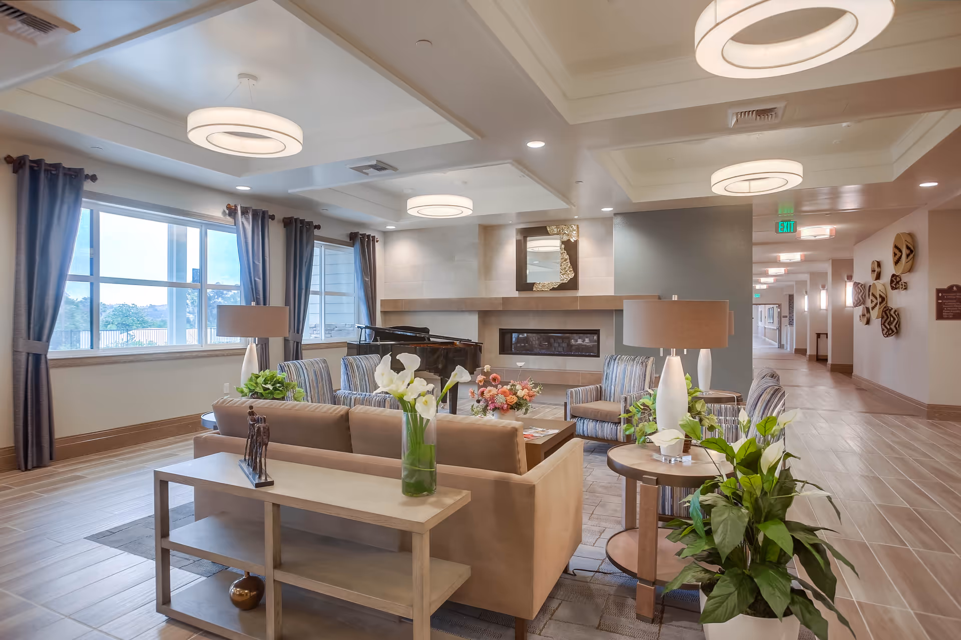 A spacious and well-lit living room area in a senior living facility with beige sofas, striped armchairs, a coffee table with floral arrangements, a modern fireplace, large windows with curtains, and ceiling lights. There is a hallway visible to the right with wall decorations and exit signs.