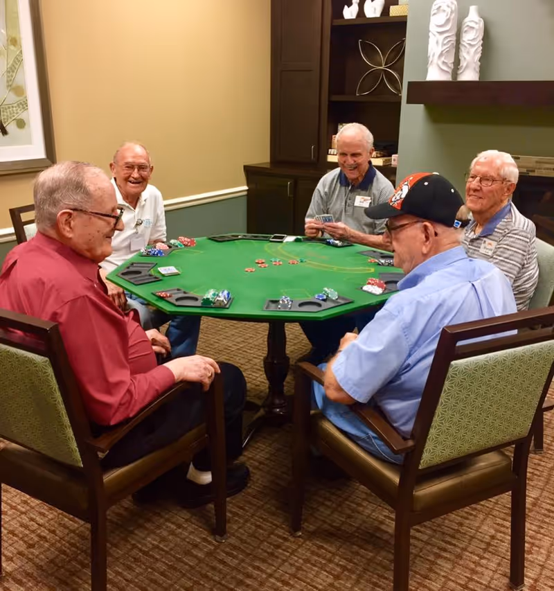 Five elderly men sitting around a green poker table playing cards and poker chips in a cozy room with carpeted floor, wooden chairs, and decorative shelves in the background.