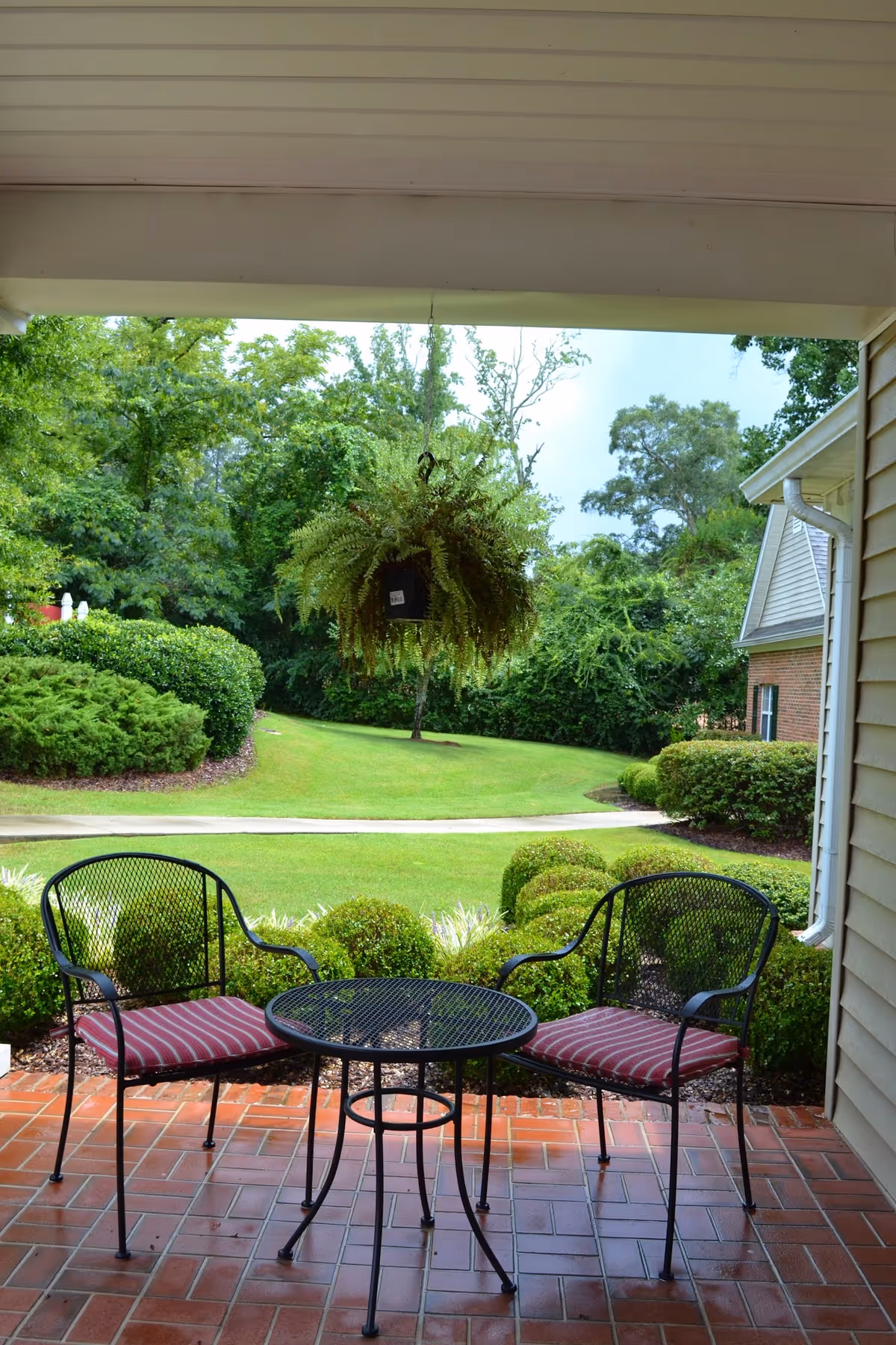 A covered patio area with two black metal chairs with red striped cushions and a matching round table. A hanging fern plant is suspended above the table. The patio overlooks a well-maintained lawn with trimmed bushes and trees in the background. Part of a building with beige siding and a white gutter is visible on the right side.