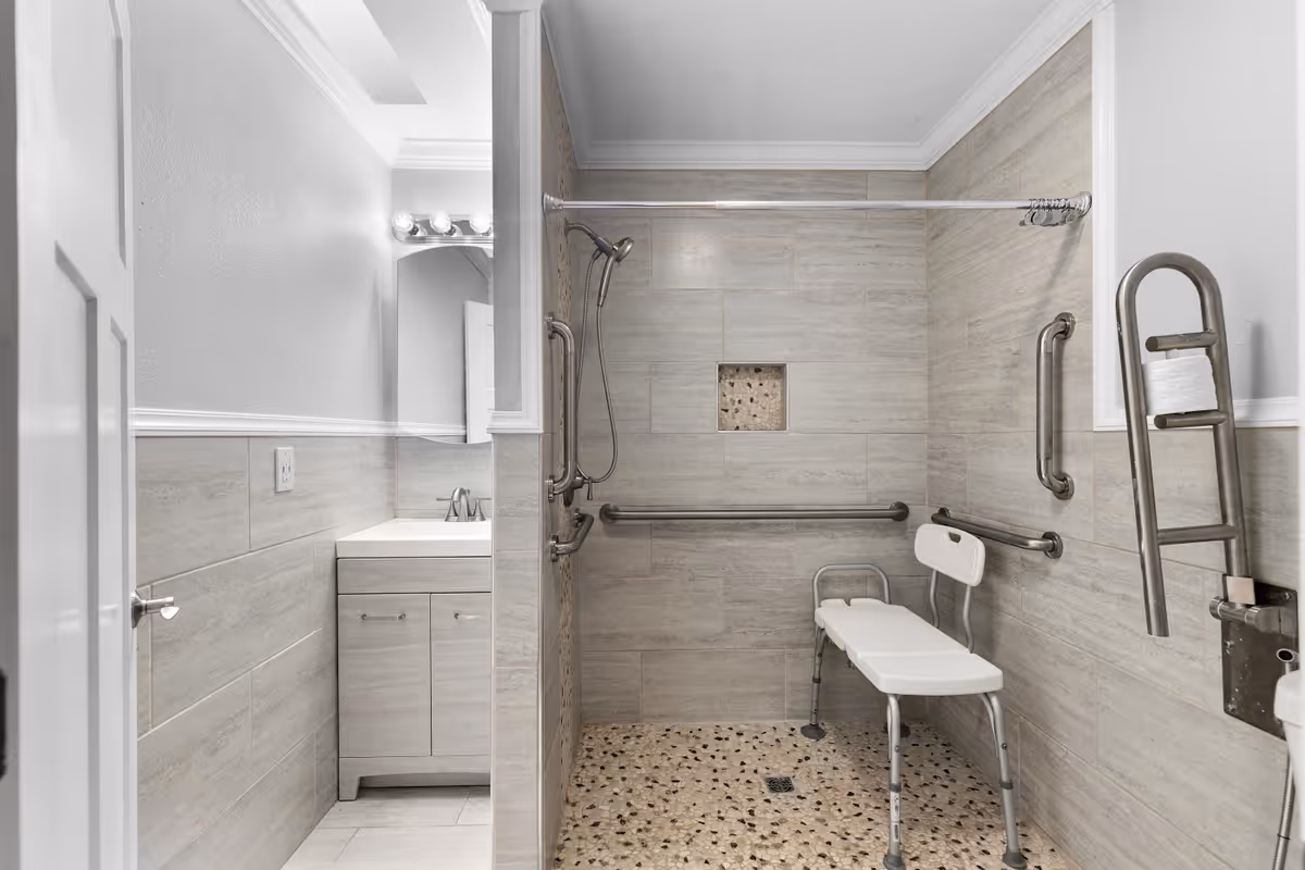 A modern accessible bathroom with a walk-in shower featuring grab bars, a handheld showerhead, and a white shower chair. The walls are tiled in light beige, and the floor has a pebble-like texture. To the left, there is a vanity with a sink, cabinet, and a mirror with lights above it.