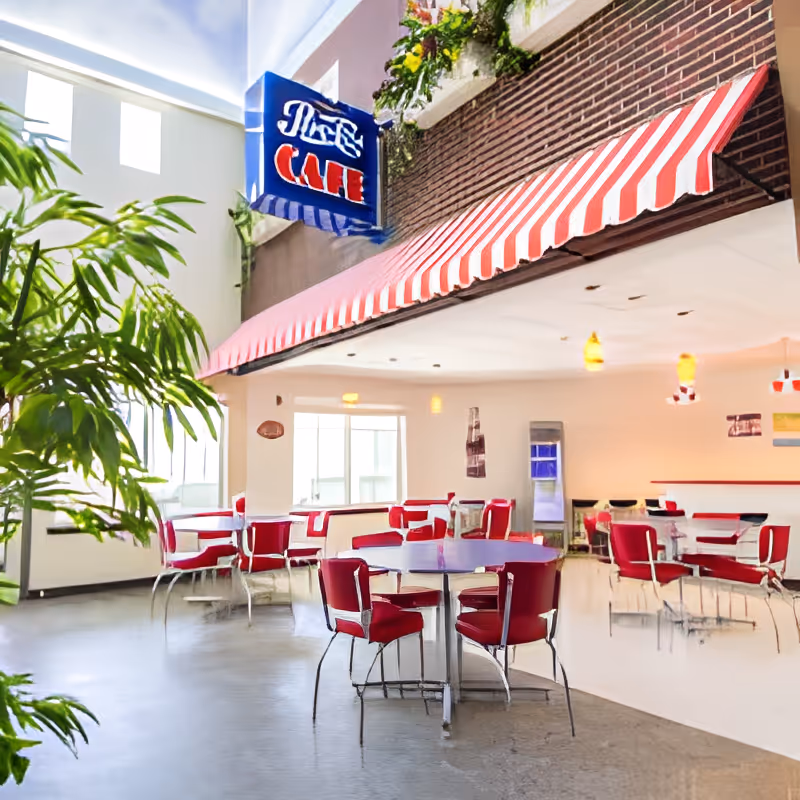 Interior view of a bright and spacious cafe area with red and white decor, featuring round tables with red chairs, a striped red and white awning, large windows letting in natural light, and green plants on the left side.